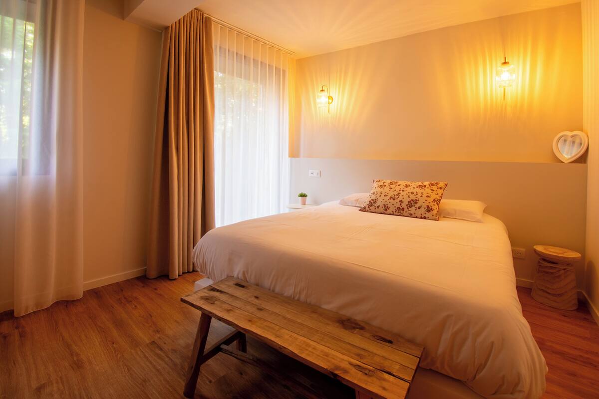 Une chambre à coucher avec un lit, un banc en bois, des rideaux, une lampe fixée au mur et une décoration en forme de cœur.