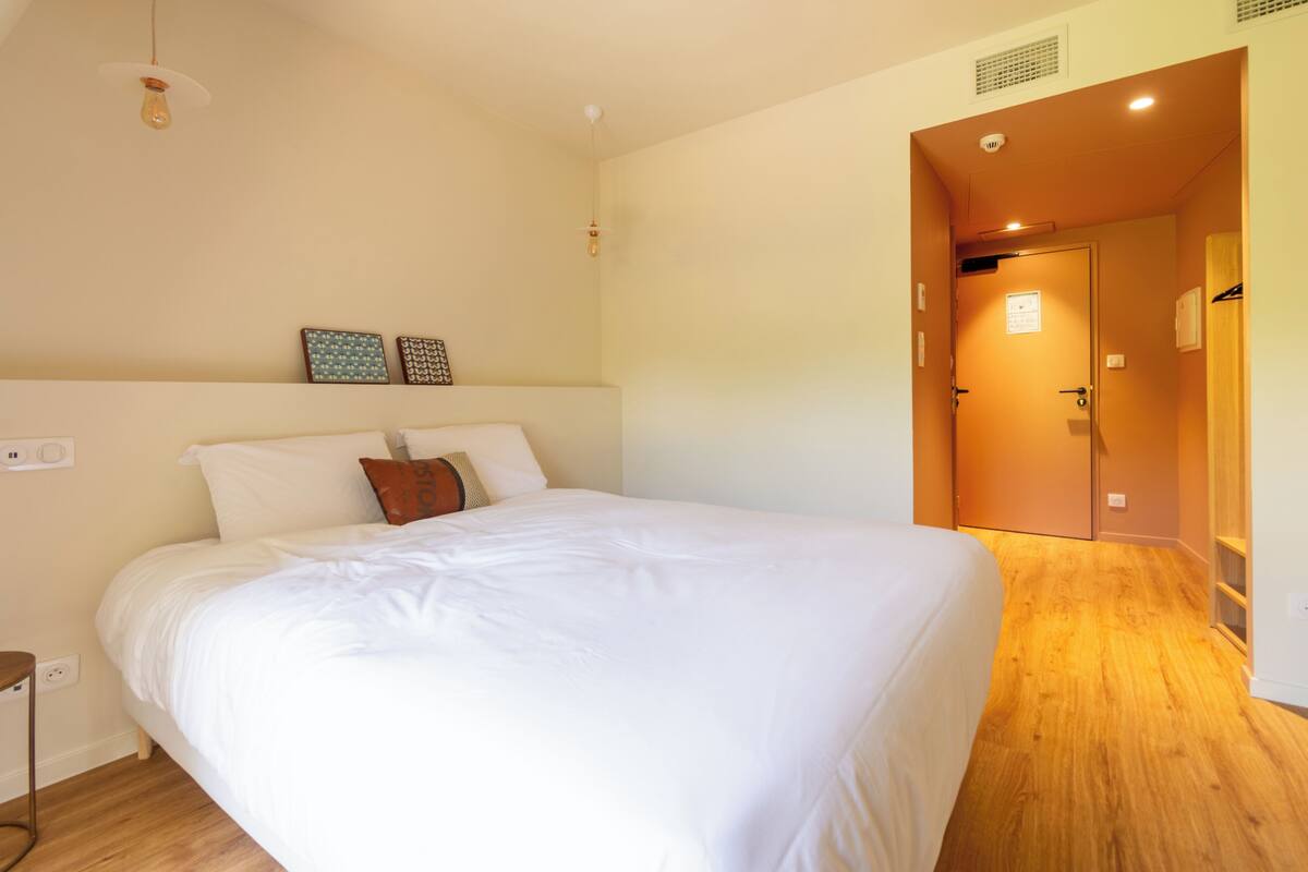 Une chambre avec un grand lit, un plancher en bois, une porte donnant sur une autre pièce et un luminaire au plafond.