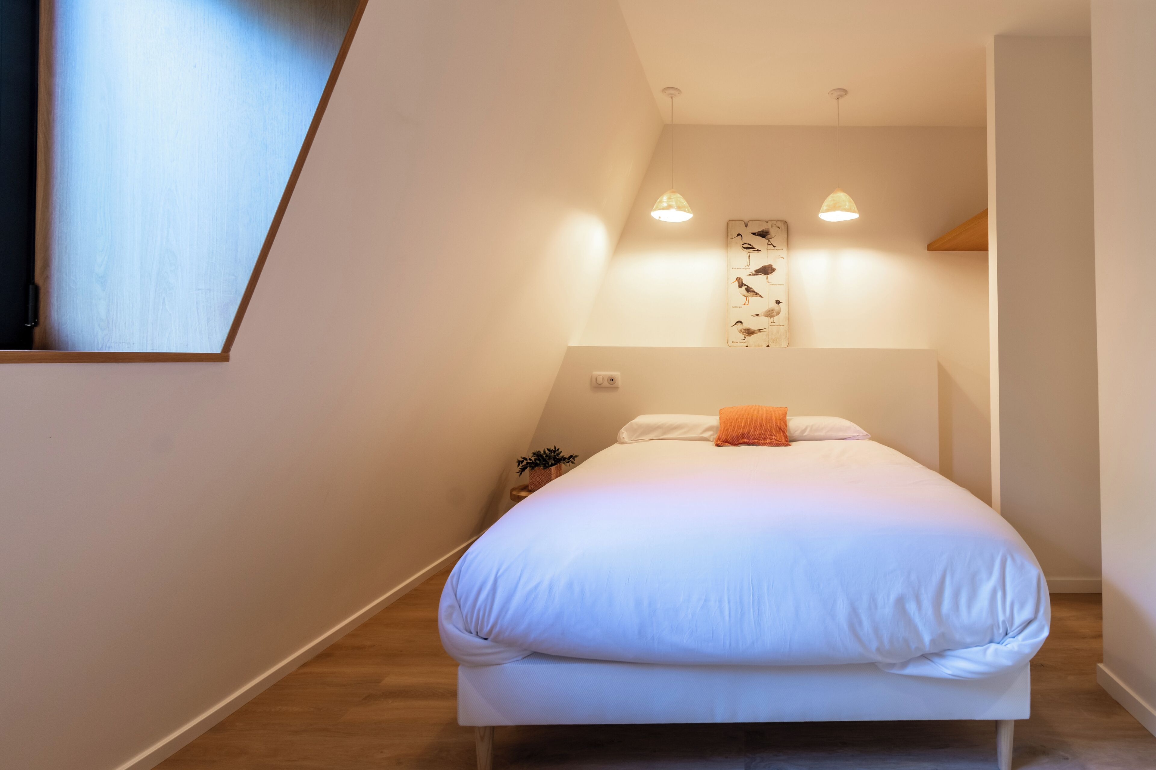 Un dormitorio con una cama, dos lámparas colgantes, una obra de arte en la pared y una planta pequeña.