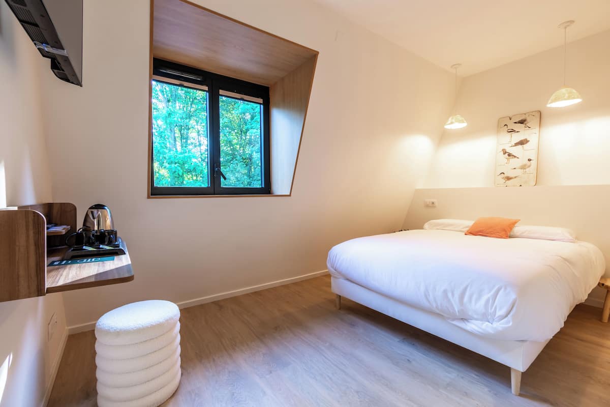 Une chambre avec un lit, une fenêtre donnant sur des arbres, une étagère avec une bouilloire et une œuvre d’art accrochée au mur.
