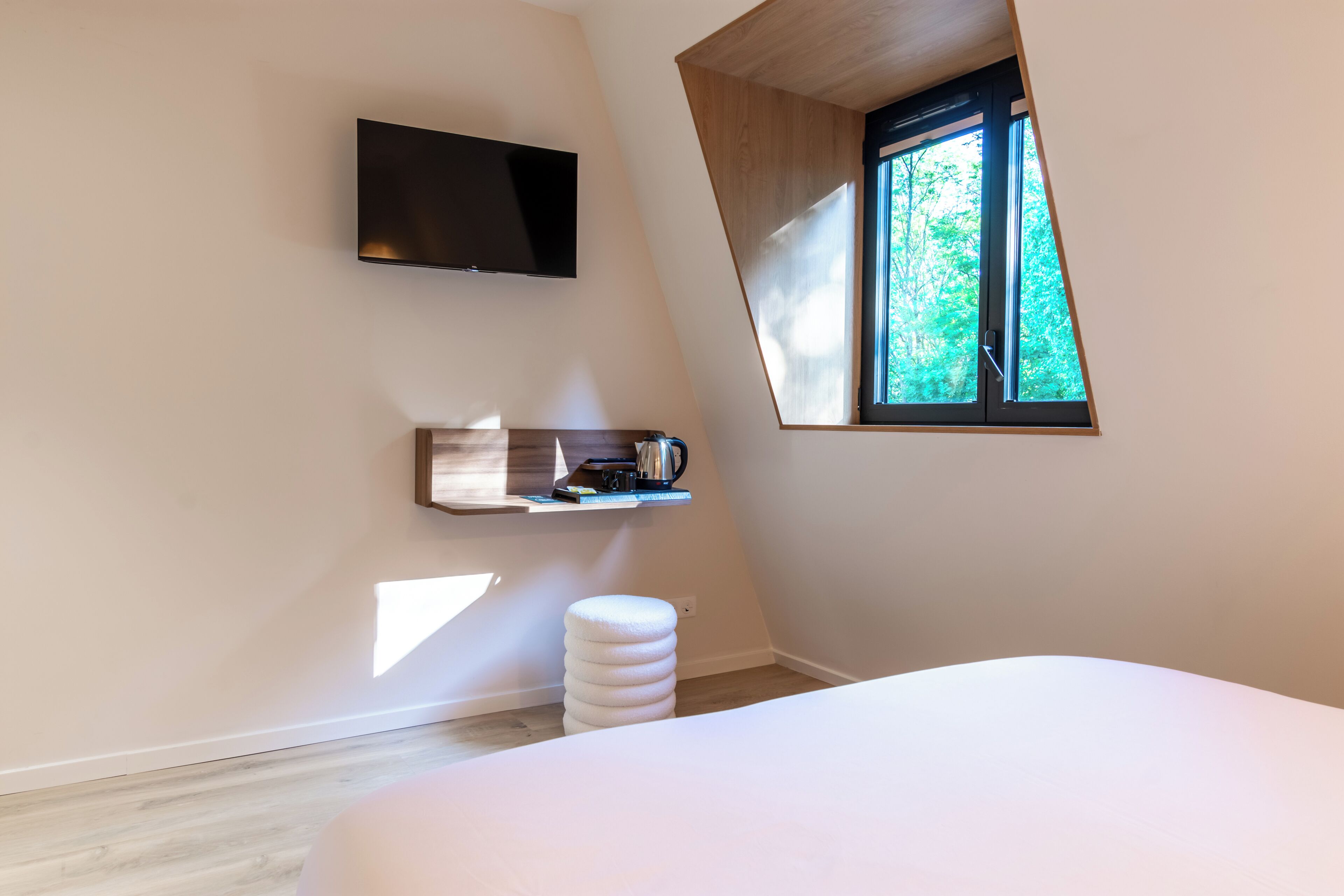 A bedroom with a bed, a TV mounted on the wall, a window, and a small shelf with items.