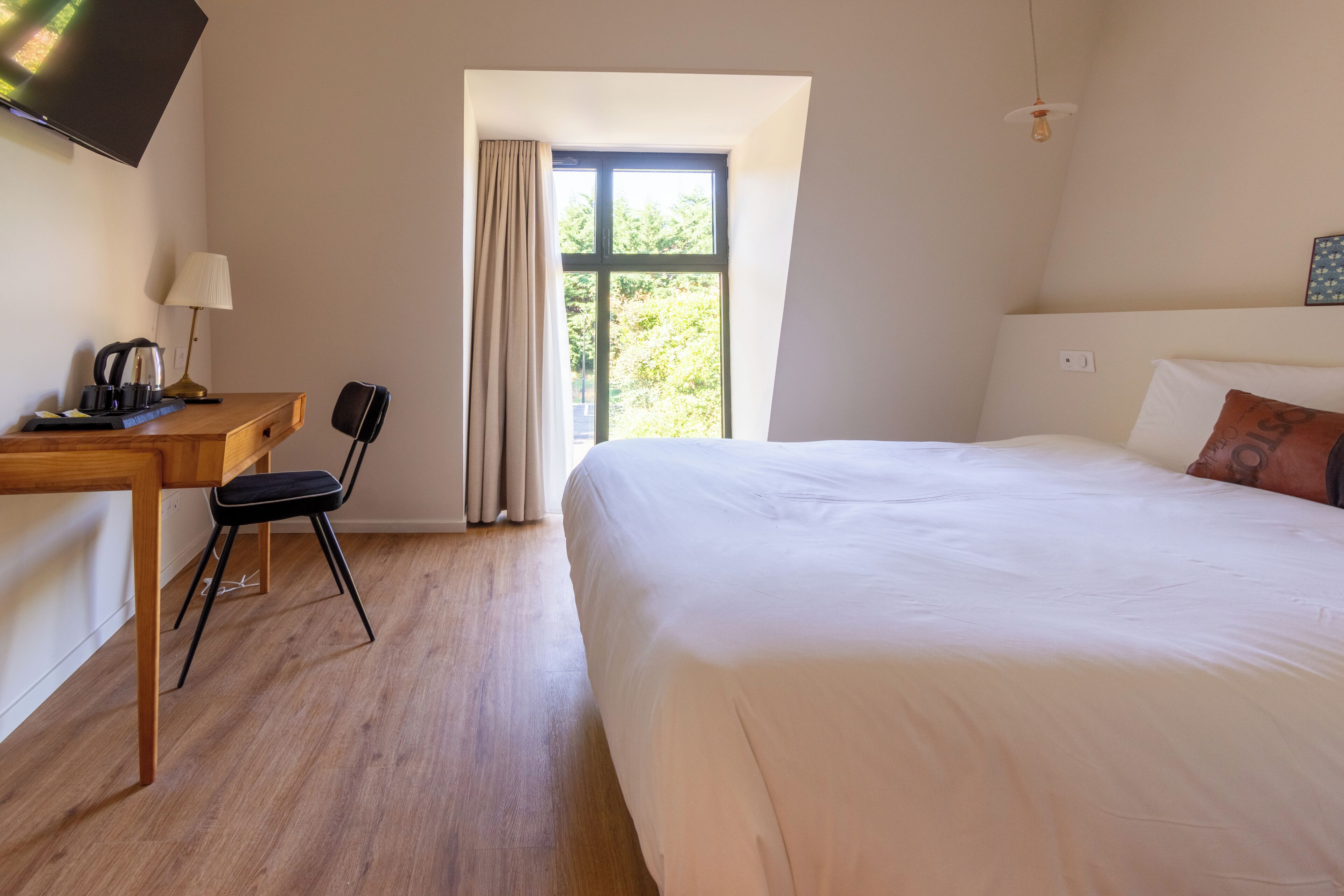 Quarto de hotel com cama, escrivaninha com luminária, cadeira, televisão fixada na parede e janela com cortinas.