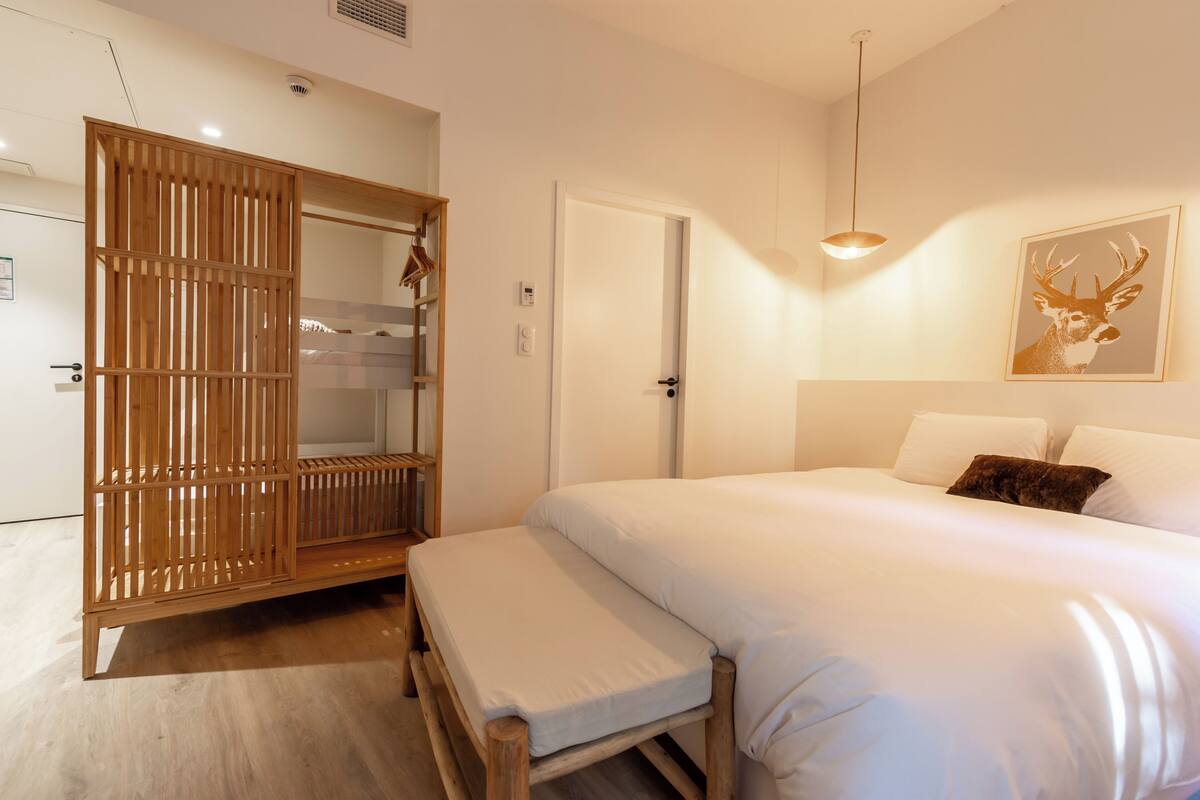 Une chambre à coucher comprenant un lit, une armoire en bois, un banc, une porte et un luminaire fixé au mur.