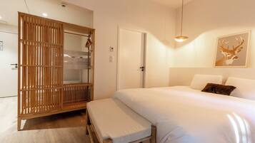 A bedroom with a bed, a wooden wardrobe, a bench, a door, and a wall-mounted light fixture.