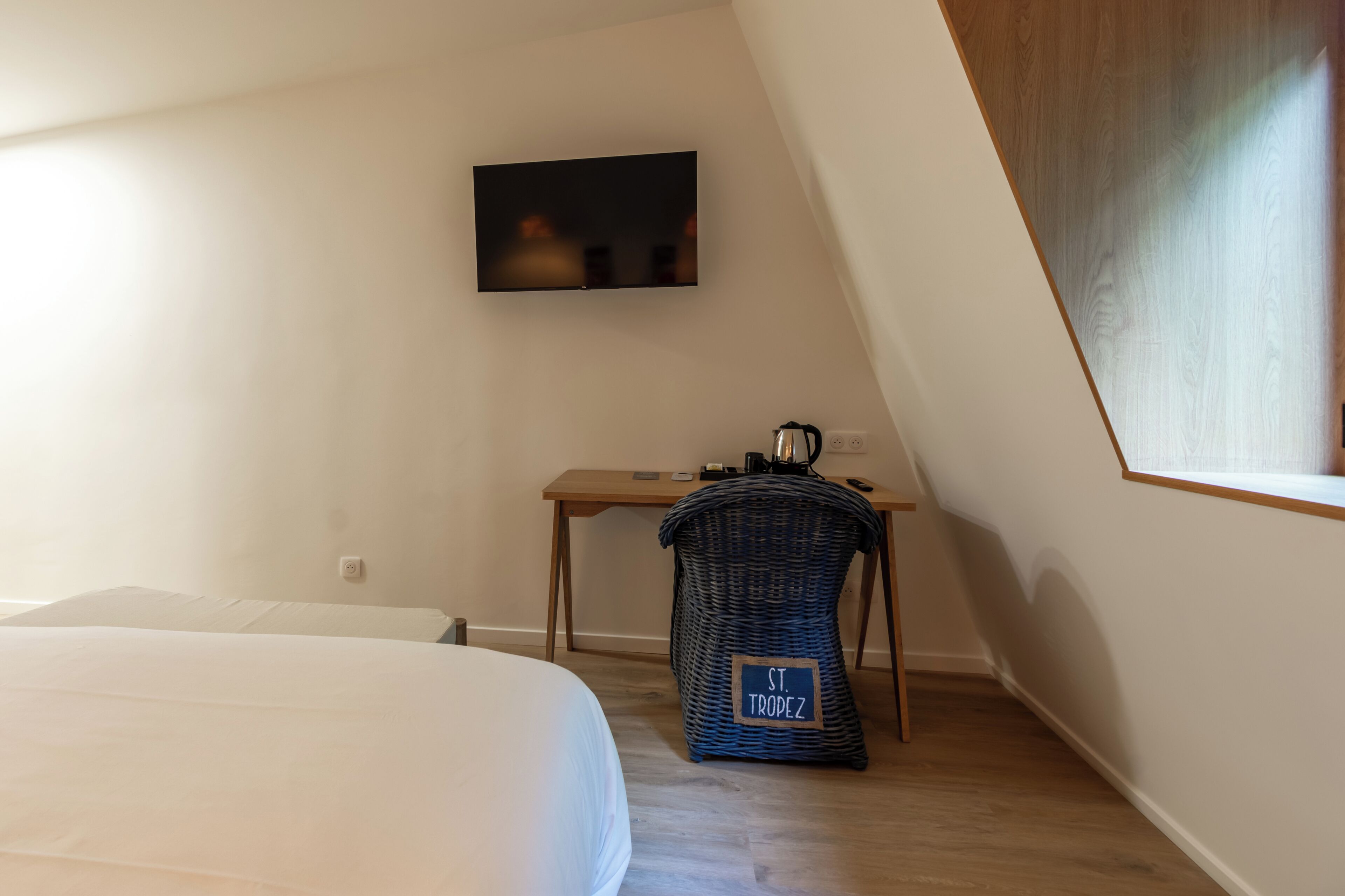 Un dormitorio con una cama, un escritorio, un televisor y una cesta de mimbre con la etiqueta 'ST. TROPEZ'.
