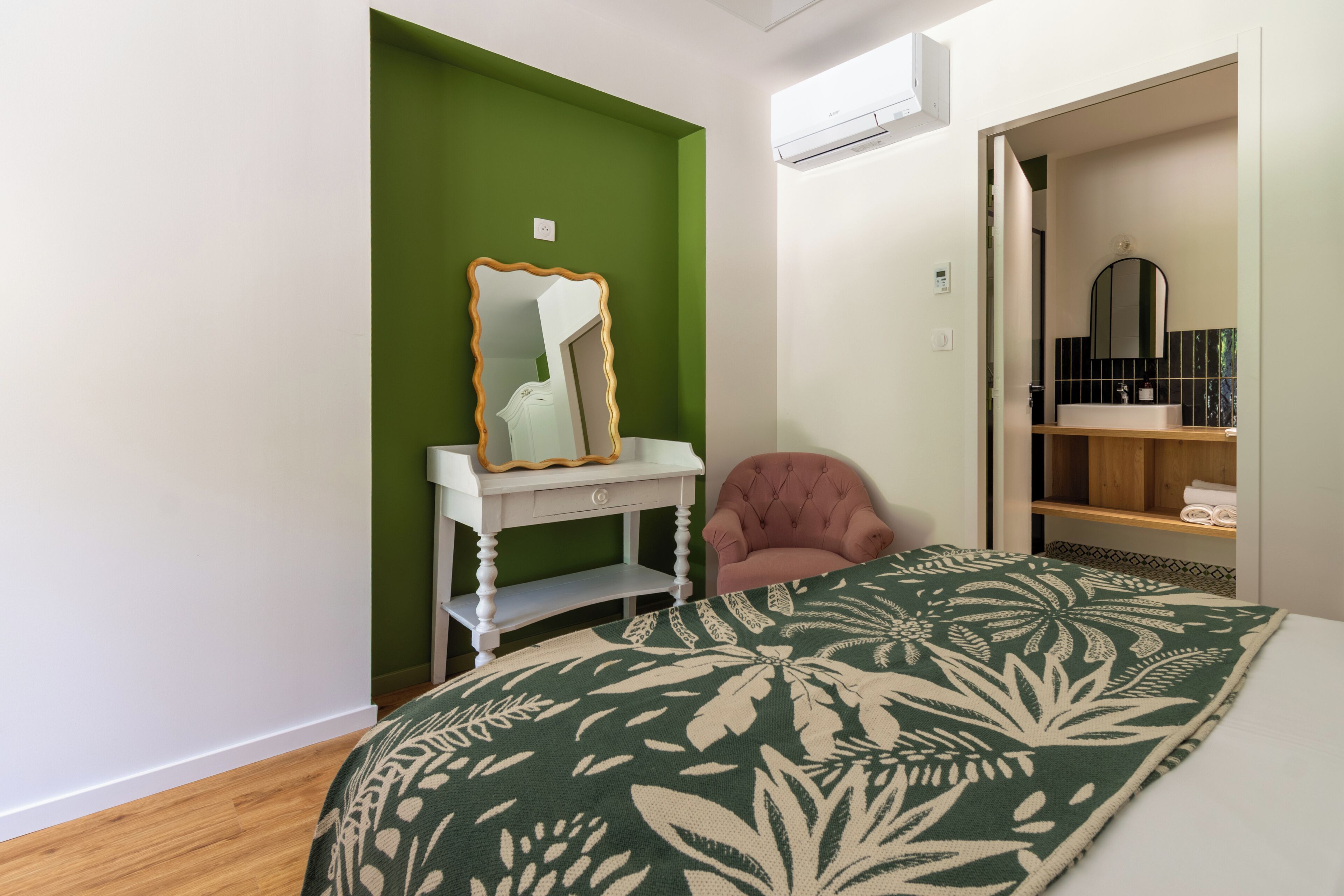Un dormitorio con cama, cómoda, espejo, silla y un baño visible a través de una puerta abierta.