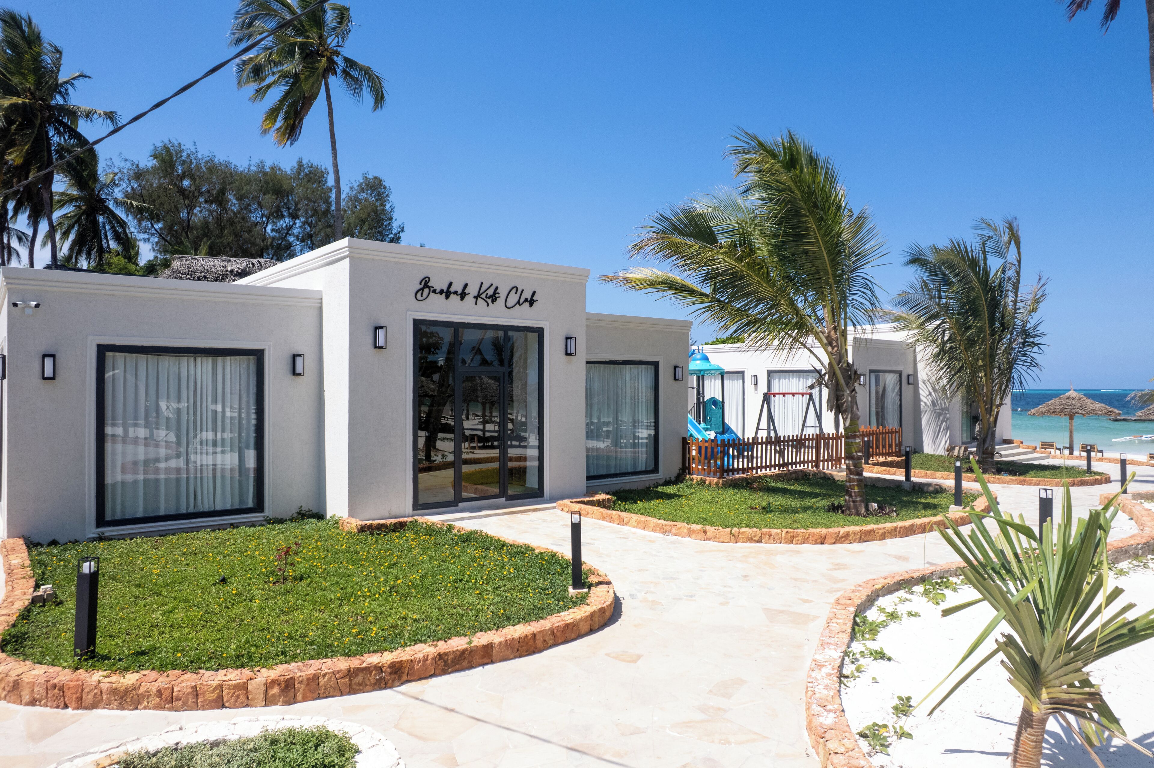 A beachfront property with a 'Beach Club' sign, palm trees, and a clear view of the ocean.