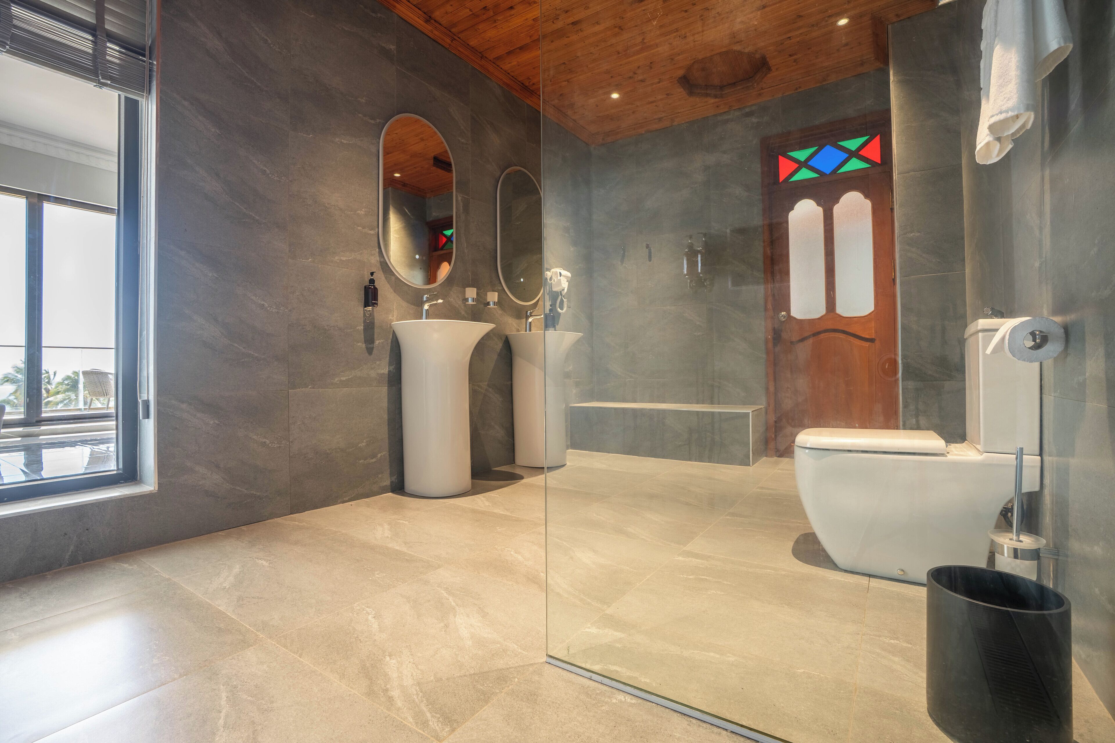 A modern bathroom with a glass shower enclosure, a large mirror, two sinks, and a wooden door with stained glass.