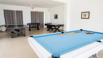 A pool table with a blue felt surface, a foosball table, and a ping pong table in a room with white walls and a ceiling fan.