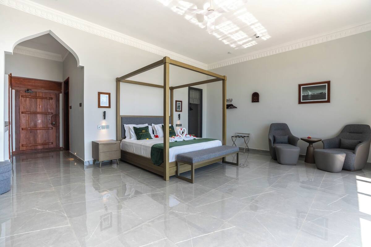 Een moderne slaapkamer met een hemelbed, een bankje, twee fauteuils, een klein tafeltje en een deur naar een andere kamer.