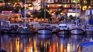 Un puerto deportivo con barcos amarrados, rodeado de edificios con letreros iluminados, incluyendo un supermercado y un restaurante.