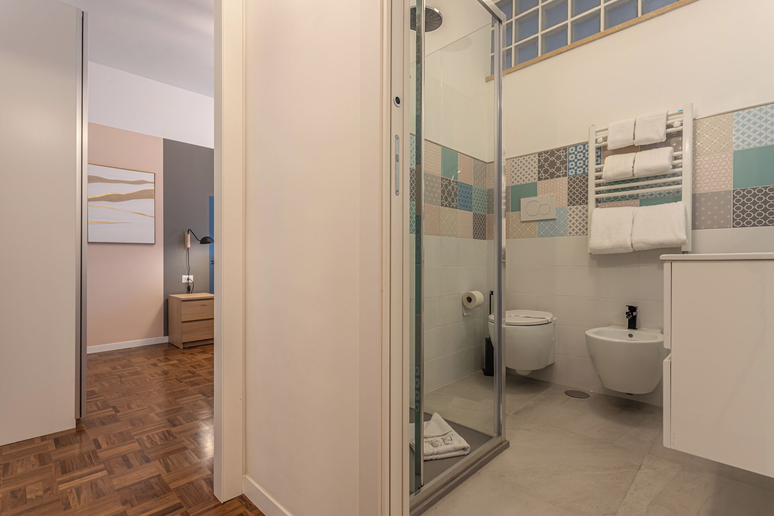 Een moderne badkamer met een glazen douchecabine, toilet en een douchetoilet. De vloer is van hout, en de wanden zijn betegeld met een geometrisch patroon.