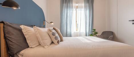Quarto com cama, escrivaninha, cadeira, luminária e janela com cortinas.