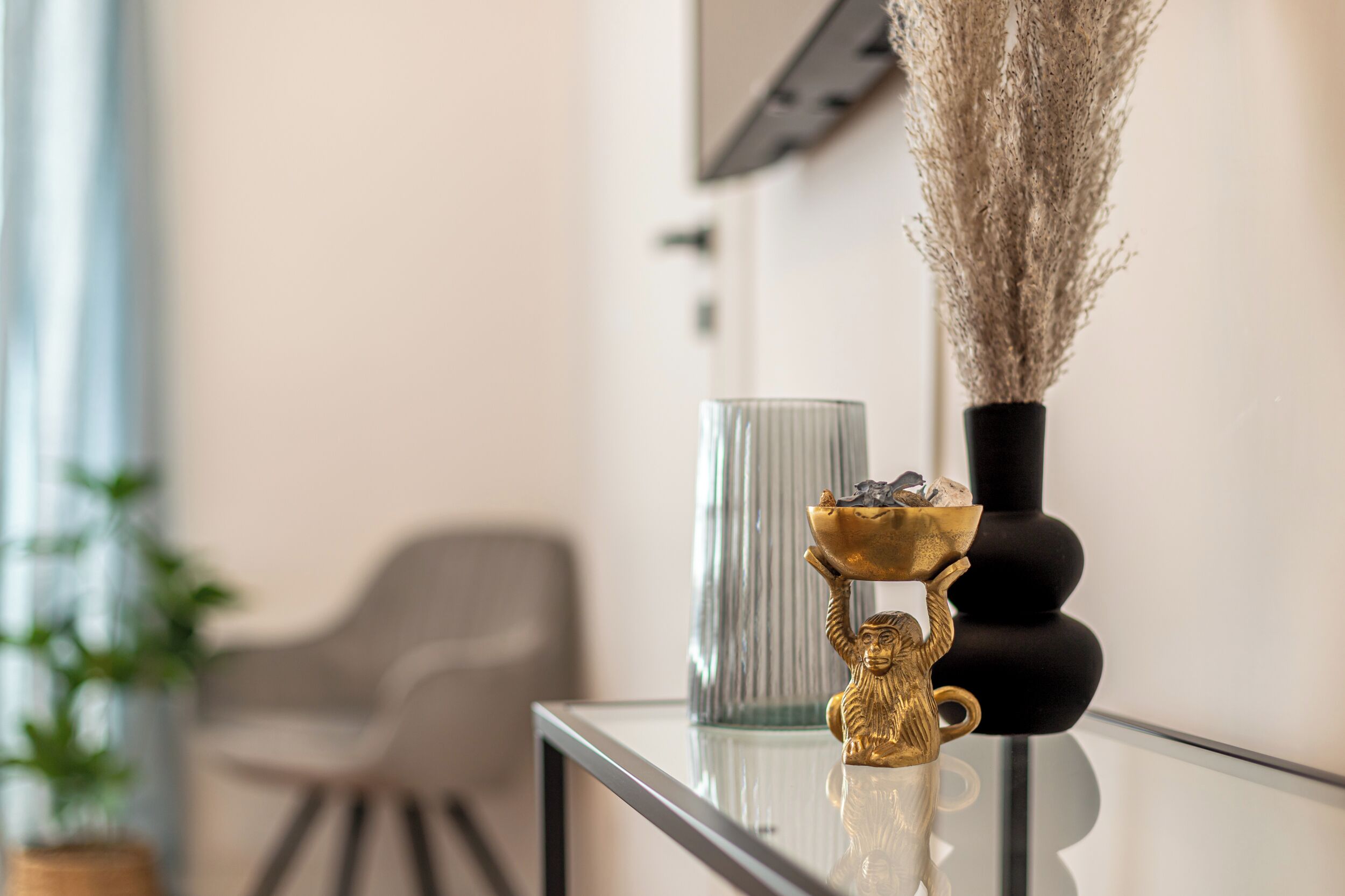 A modern interior with a glass table displaying decorative items, a grey chair, and a plant in the background.
