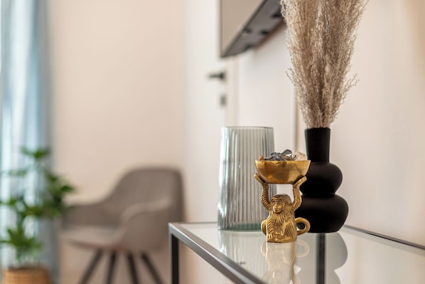 A modern interior with a glass table displaying decorative items, a grey chair, and a plant in the background.