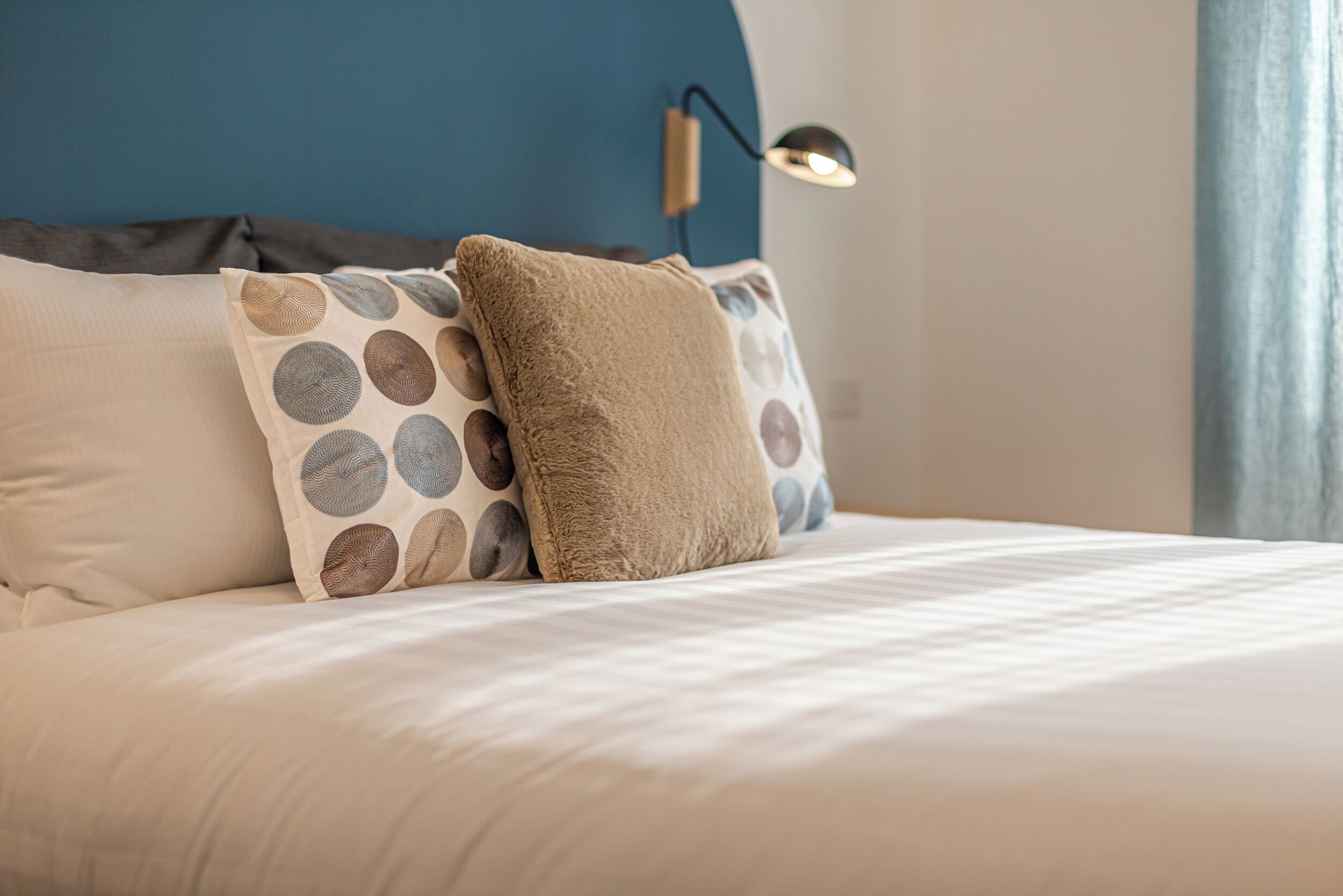 A neatly made bed with multiple pillows, a blue wall, and a wall-mounted lamp.