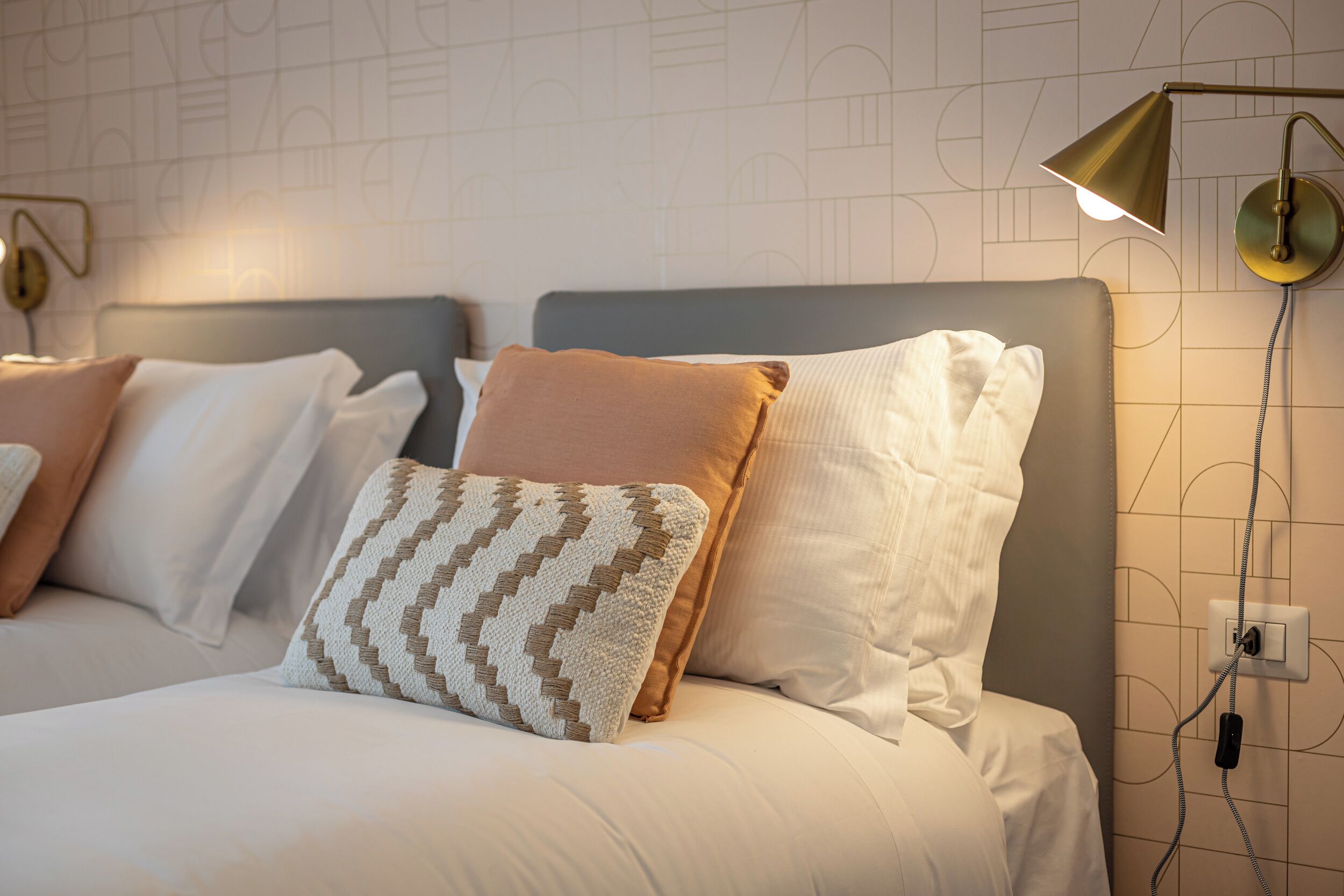 A neatly made bed with multiple pillows, a bedside lamp, and a wall socket.
