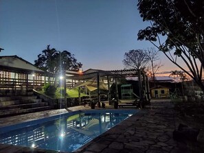 A swimming pool with a stone pathway, a covered seating area, and a building with a staircase in the background. - vereda bike park hotel (São João del Rei)