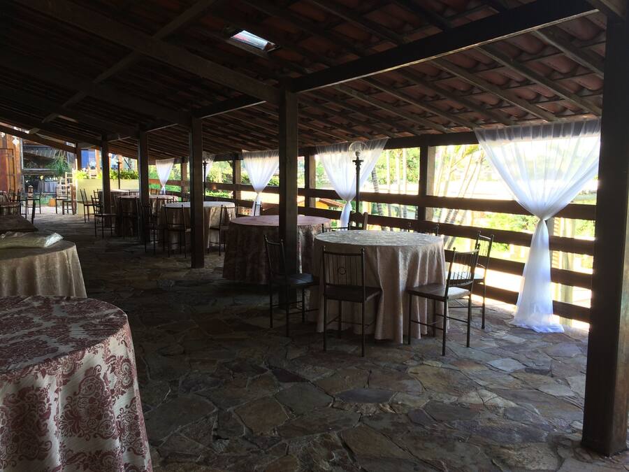 Un patio al aire libre con suelo de piedra, mesas redondas con sillas y una estructura de techo de madera.