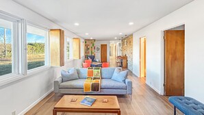 A spacious living room with a grey sofa, a wooden coffee table, and a view of a garden through large windows. - Retro Retreat Central Location Garage (Grand Junction)