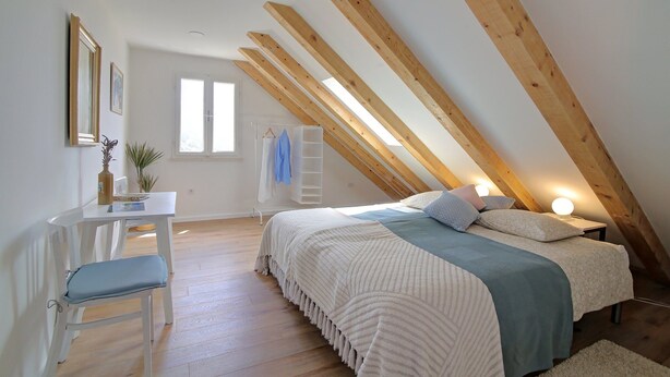 Ein Schlafzimmer mit schräg angeschnittenem Dach, einem Bett mit Kissen, einem Nachttisch, einem Stuhl, einem kleinen Tisch mit Pflanze und einem Fenster.