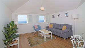 Ein modernes Wohnzimmer mit grauem Sofa, einem weißen Couchtisch, einem blauen Sessel und einer Stehlampe. Es gibt Fenster mit weißen Rahmen und Blick auf den Himmel.