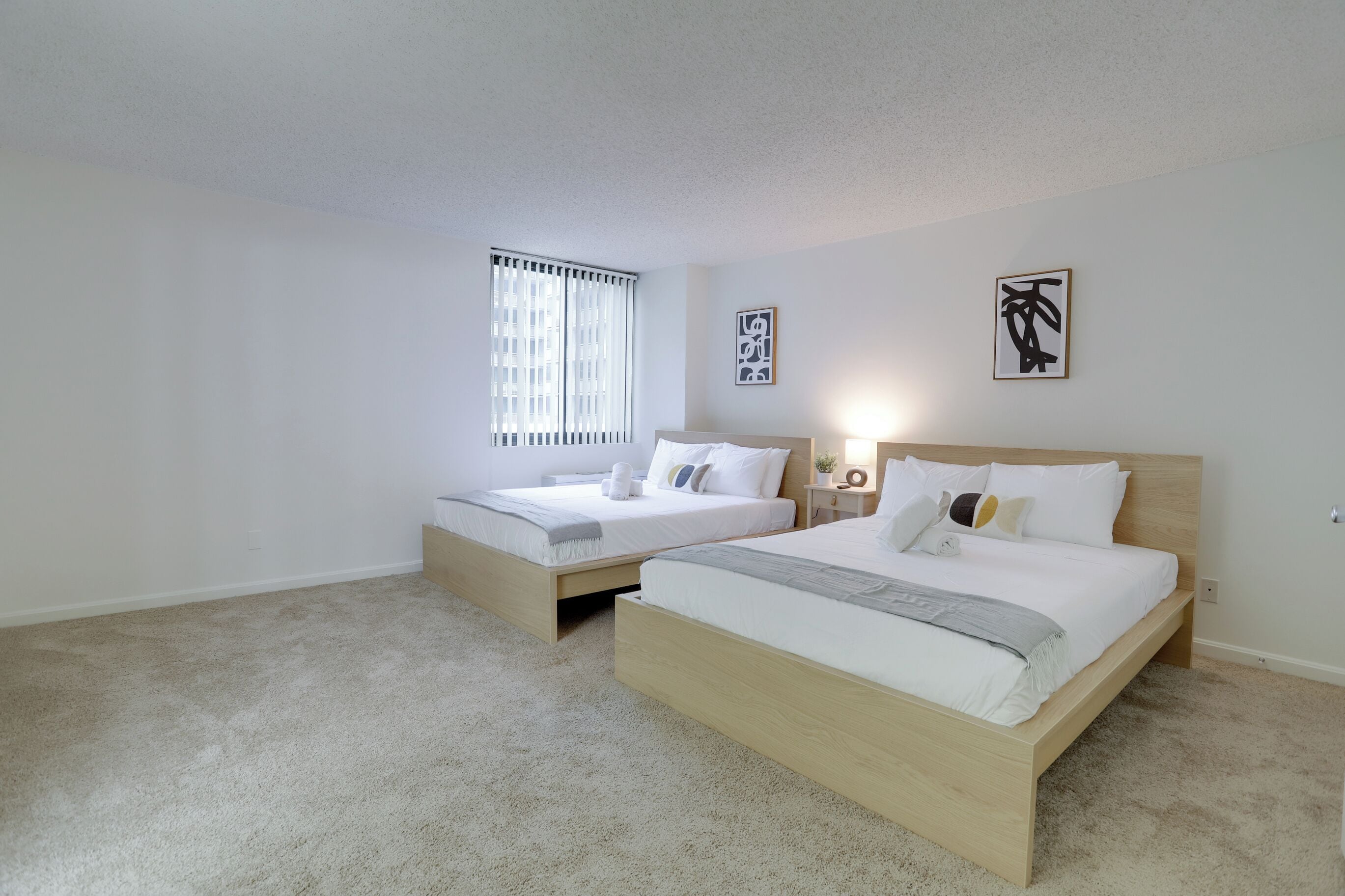 A bedroom with two single beds, a window with blinds, and two wall-mounted artworks.