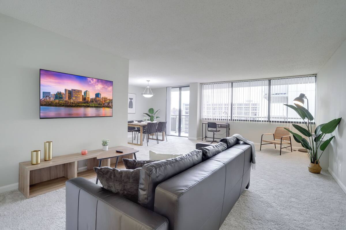 A modern living room with a grey sofa, a dining area with a table and chairs, and a large window with blinds.
