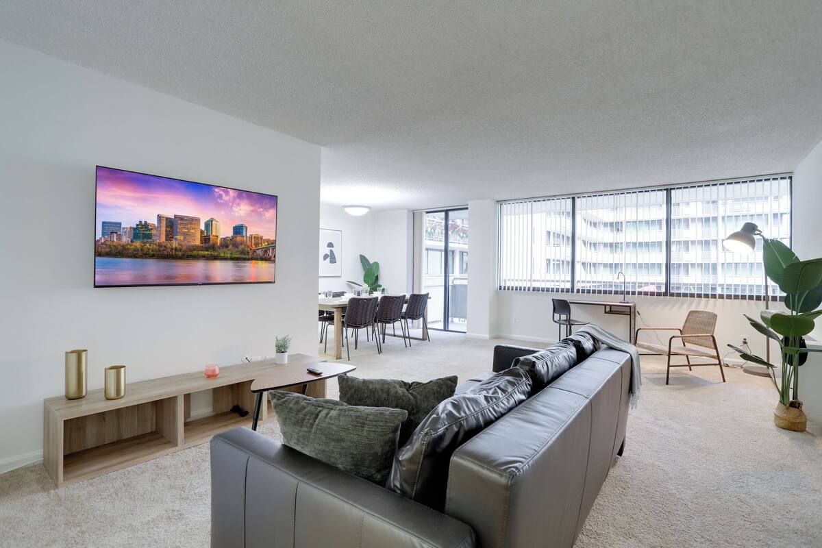 A modern living room with a grey sofa, a dining area with a table and chairs, and a large window with blinds.