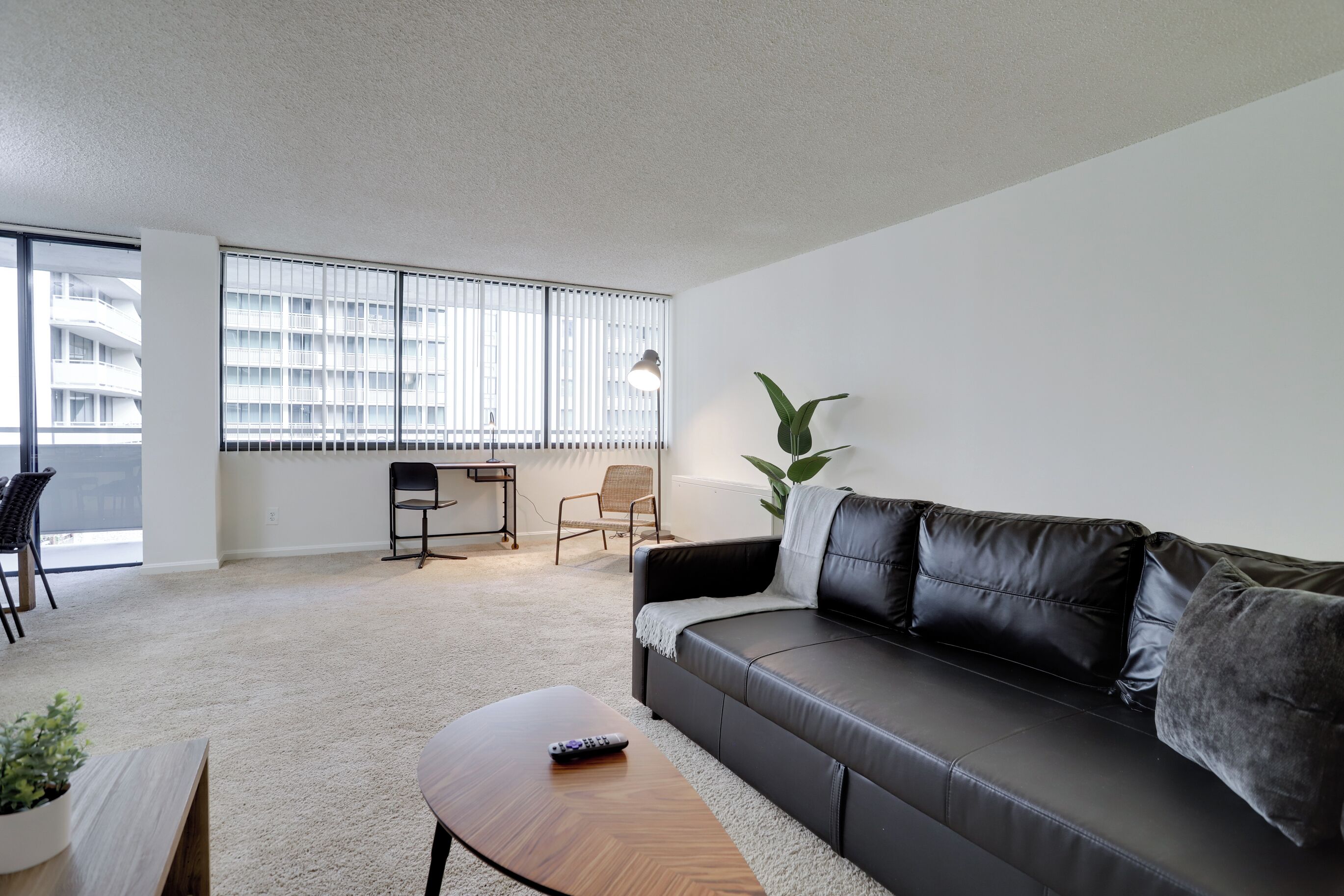 A modern living room with a black leather sofa, a wooden coffee table, and a large window with blinds.