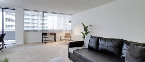 A modern living room with a black leather sofa, a wooden coffee table, and a large window with blinds.