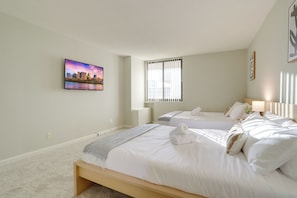 A hotel room with two beds, a cityscape painting, a window with blinds, and a bedside lamp. - Arlington Apartments near Ballston - We Host (Arlington)