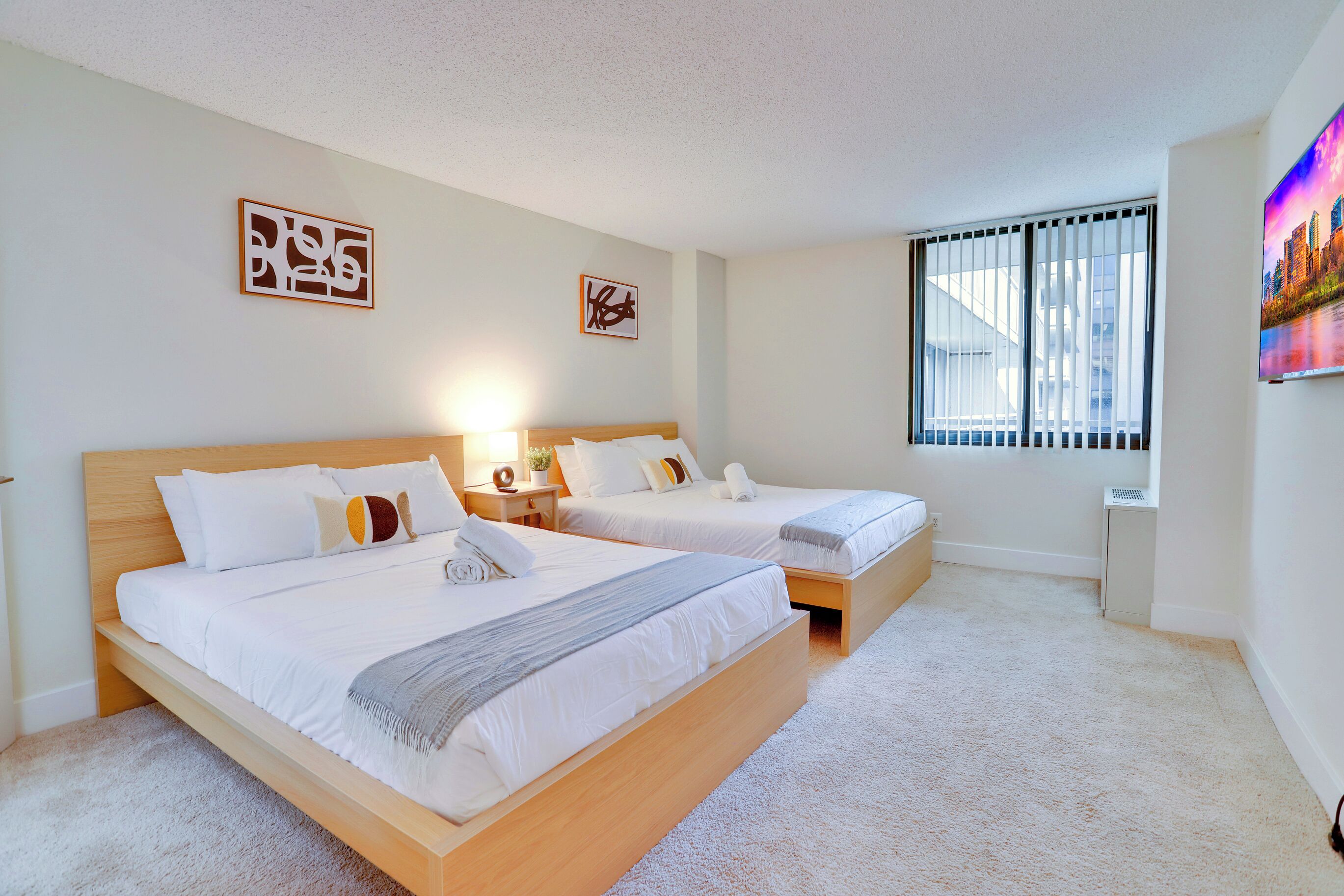 Een hotelkamer met twee bedden, een groot raam met zonwering en kunstwerken aan de muur.