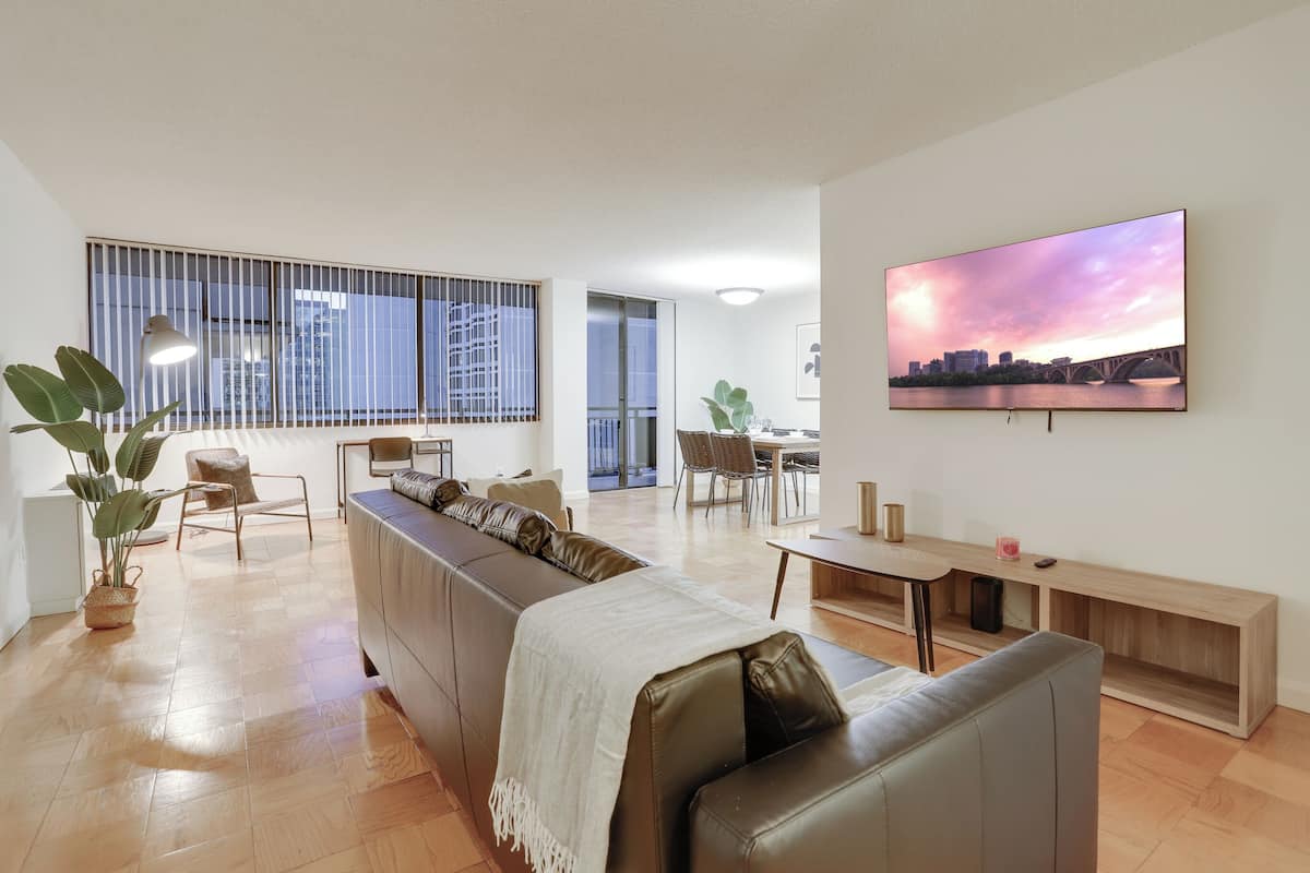 A modern living room with a long brown sofa, a dining area with a table and chairs, and a large TV on the wall.