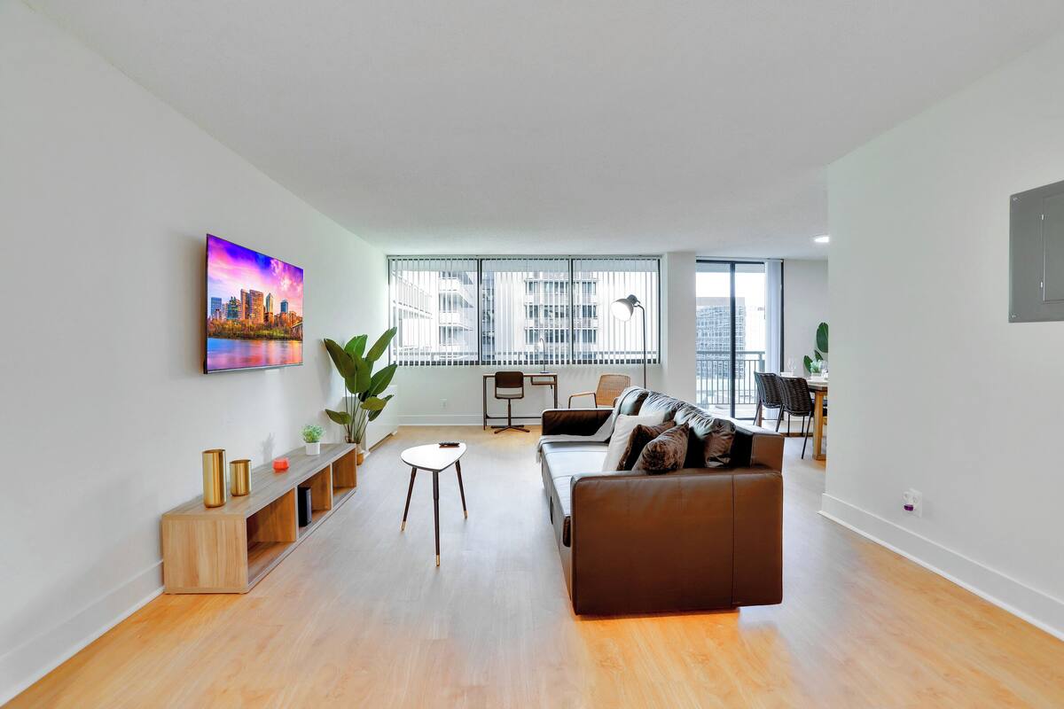 A modern living room with a brown sofa, a coffee table, and a TV mounted on the wall. There is a large window with blinds, a plant, and a small table with chairs.