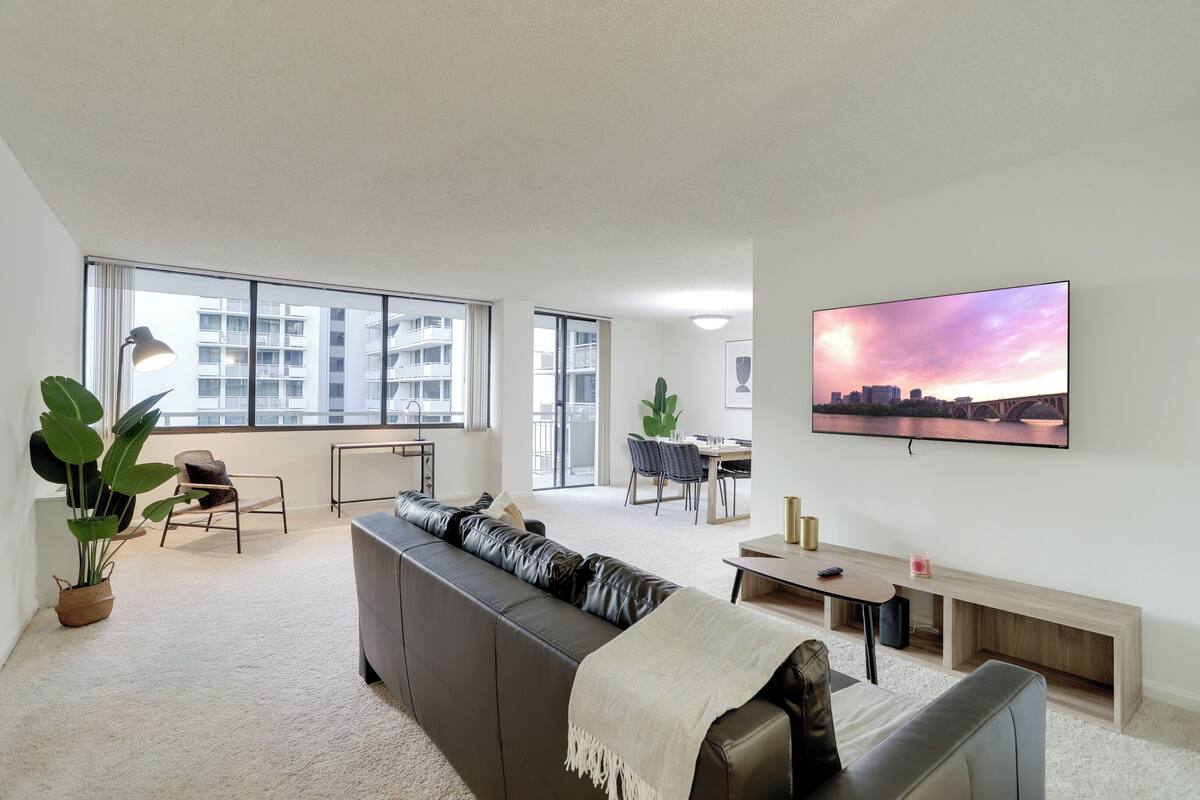 A modern living room with a large window, a sofa, a dining area, and a flat-screen TV mounted on the wall.