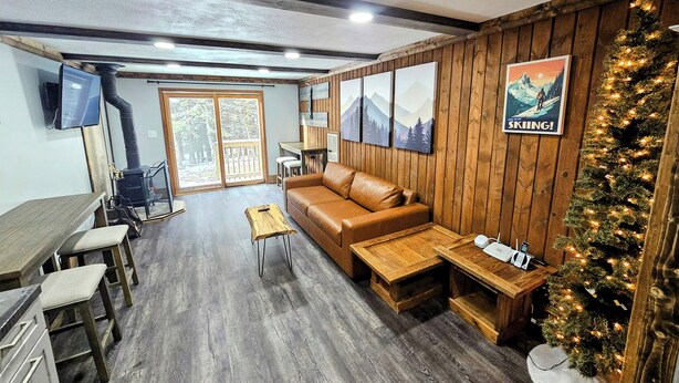 A cozy living room with a wooden wall, a leather sofa, a small wooden table, and a Christmas tree. - Shamrock 73 - 2br/2ba - Pet Friendly - Fully Renovated! (Snowshoe)