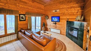 Ein Blockhauss Innenraum mit Ledersofa, Kamin, Fernseher und Blick ins Freie.