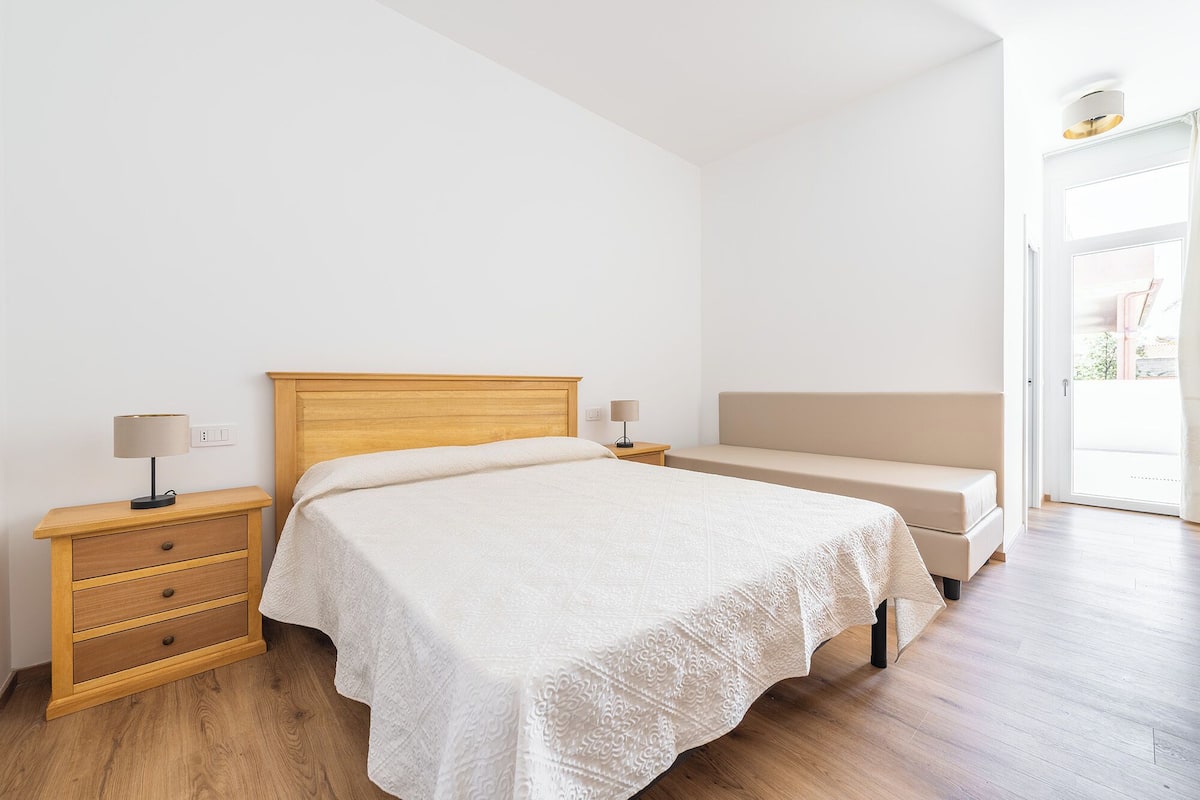 Un dormitorio con una cama de madera, una mesita de noche con lámpara, un banco y vistas al exterior.