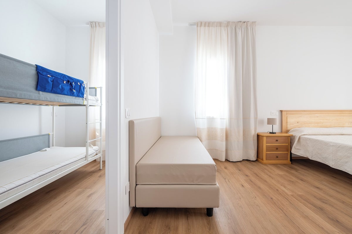 Habitación de hotel con literas, una cama individual, una mesita de noche con lámpara y suelo de madera.