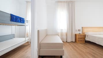 A hotel room with a bunk bed, a single bed, a bedside table with a lamp, and a wooden floor.
