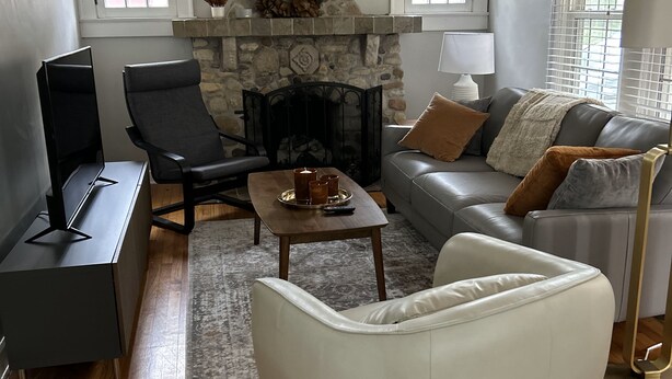 A living room with a stone fireplace, a ceiling fan, a grey sofa, a white armchair, and a wooden floor. - Charming 1925 Home In Broad Ripple - Sleeps 6, Modern Comforts & Prime Location (Indianapolis)