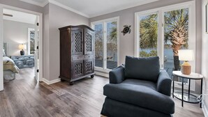 Un salón con un sillón oscuro, un aparador de madera y vistas a palmeras a través de grandes ventanales.