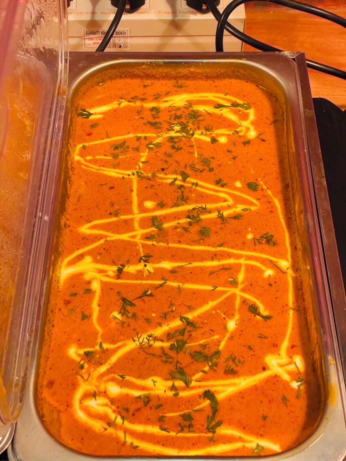 Un récipient métallique rectangulaire contenant une sauce orange crémeuse, garnie d’herbes et décorée d’un motif en forme de fines lignes jaunes.