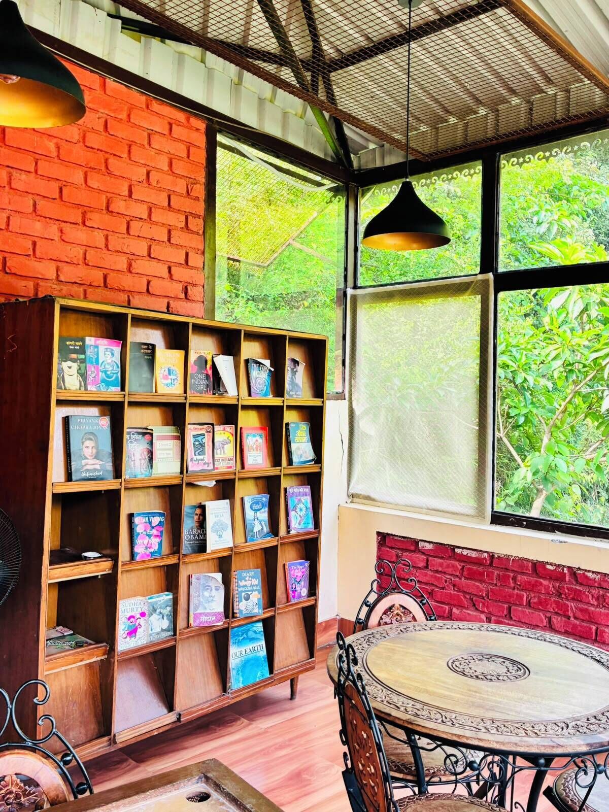 A cozy reading nook with a bookshelf, a round table, and a view of greenery through large windows.