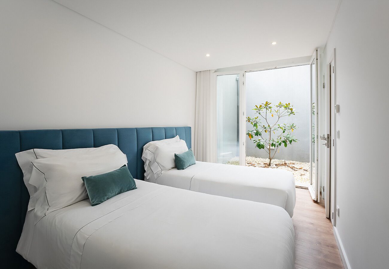 A modern bedroom with two beds, a blue headboard, and a view of a garden through an open door.