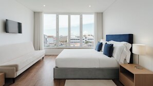Ein modernes Hotelzimmer mit einem großen Bett, einer Couch, einem Fernseher und Blick auf die Stadt durch große Fenster.
