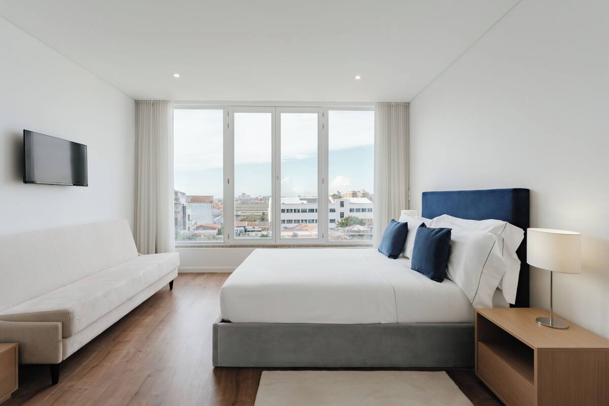 Una camera d'albergo moderna con un letto grande, un divano, una TV e vista sulla città attraverso ampie finestre.