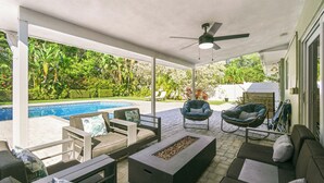 A covered outdoor patio with seating, a fire pit, and a view of a swimming pool and lush greenery. - Charming 4-Bedroom Beach Bungalow with Idyllic Pool & Backyard (Tequesta)