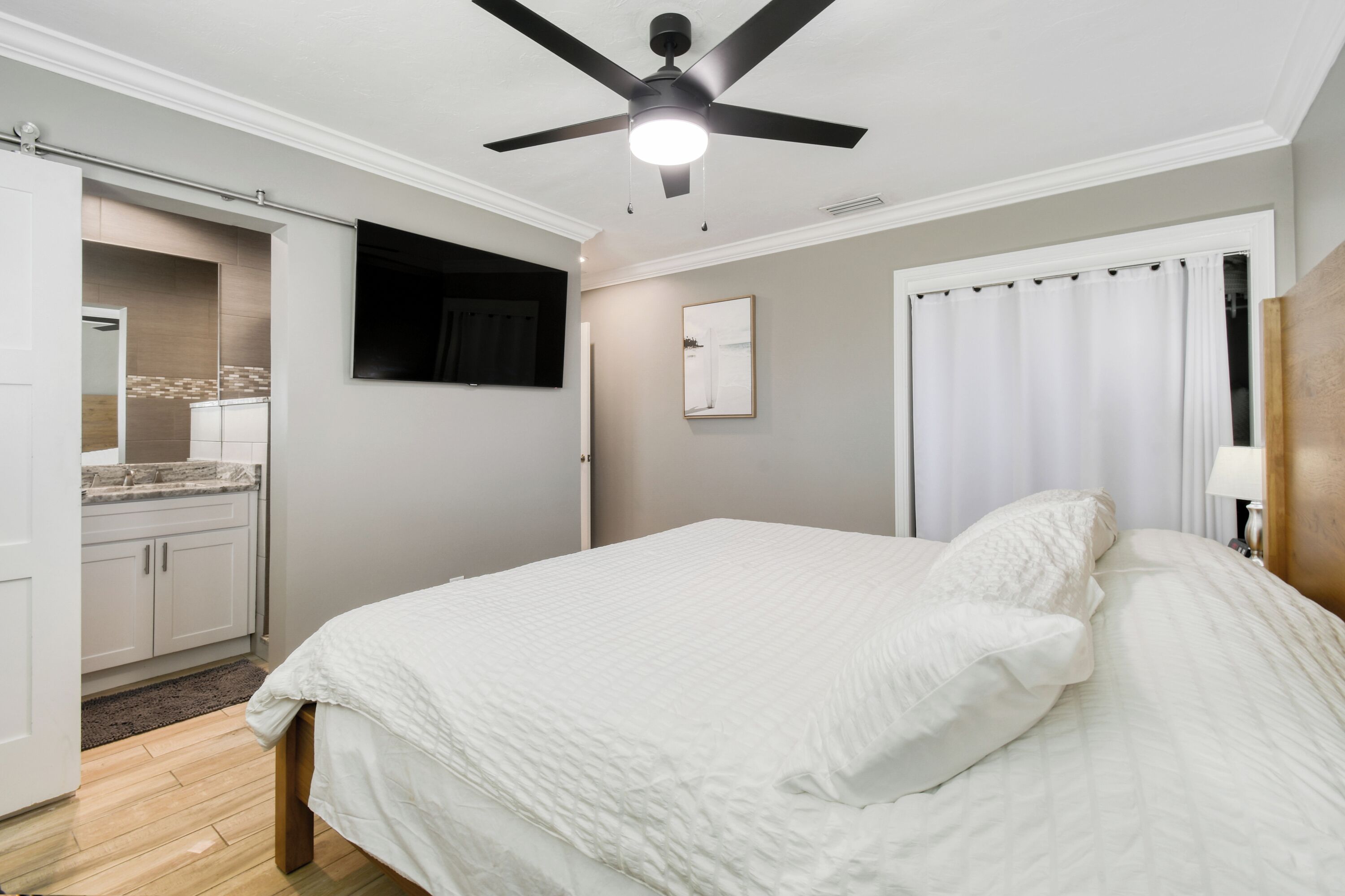 A bedroom with a bed, a ceiling fan, a window with curtains, and a bathroom with a shower and sink.